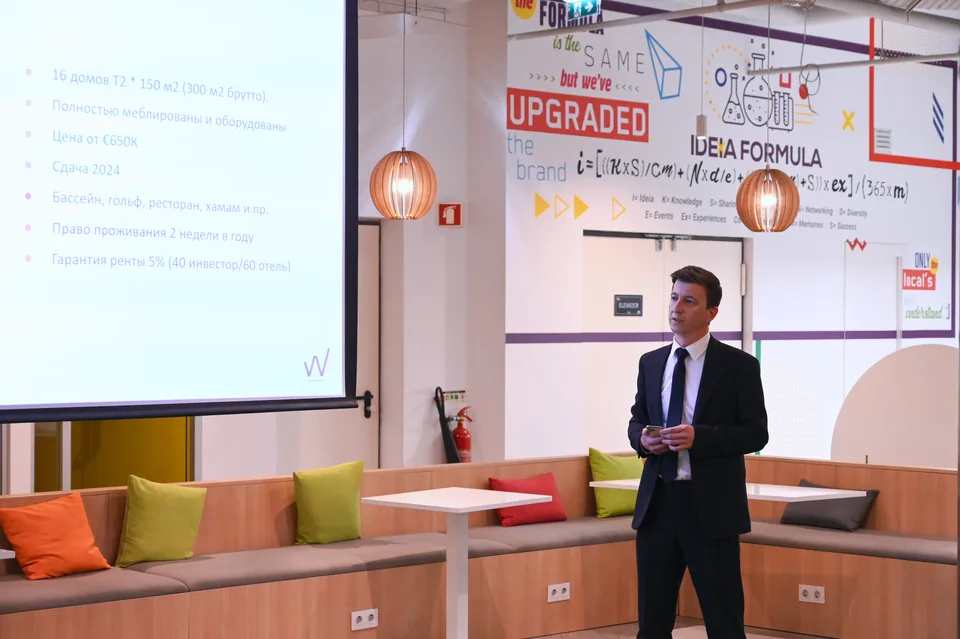 Ведущий в темном костюме стоит между экраном презентации и стеной IDEA Spaces